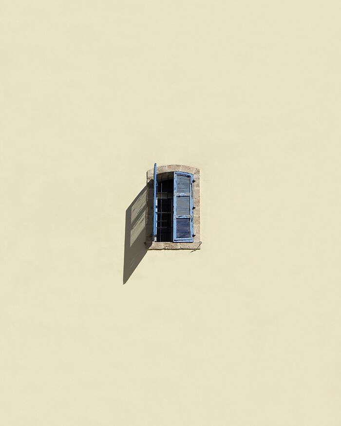Minimalist art image of a blue-shuttered window on a vast, plain wall, using negative space effectively.
