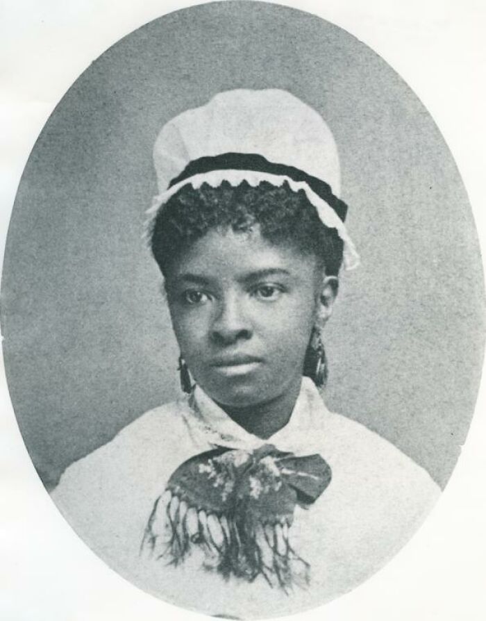 A young Black woman in a white bonnet with dark curly hair and earrings, captured in an oval portrait, highlighting women in history.