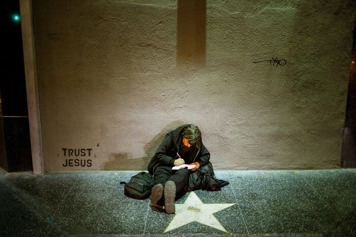 A person sits writing on the sidewalk next to a star, a perfectly timed street photo by Jeremy Paige.