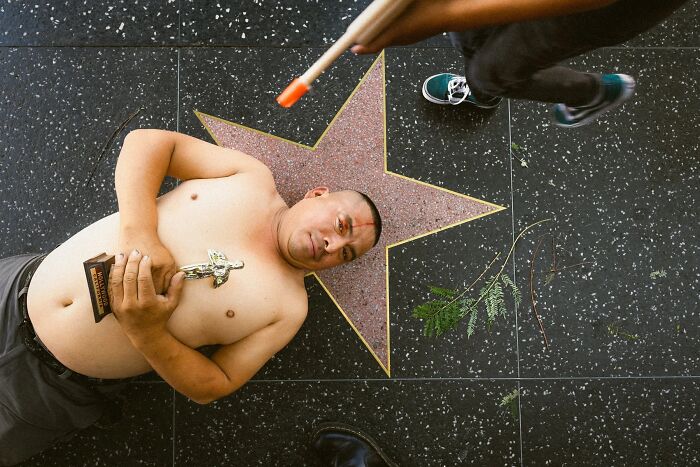 Perfectly timed street photo by Jeremy Paige showing a man holding a trophy lying on a star with a bat overhead.