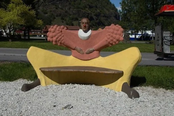 A bizarrely designed park bench shaped like a person with a red nose and large arms, another example of playground fails.