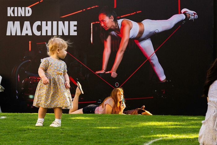 Perfectly timed street photo by Jeremy Paige showing a child and woman on grass in front of a fitness ad.