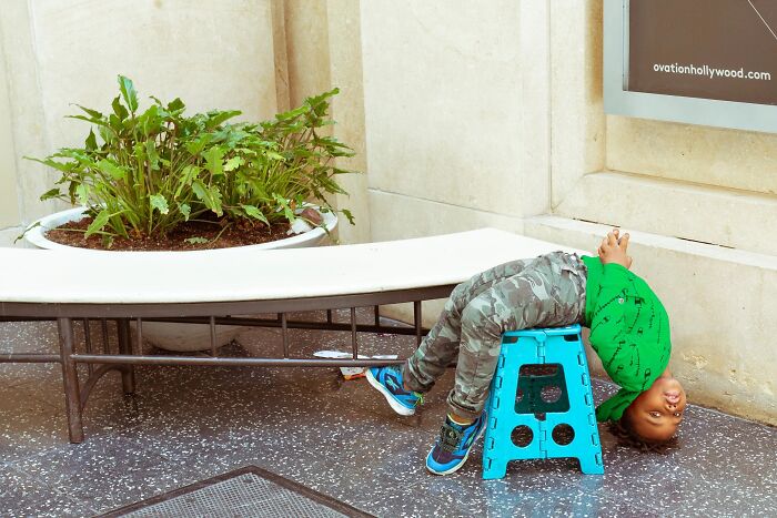 A perfectly timed street photo by Jeremy Paige shows a child upside down on a small blue stool on the sidewalk.