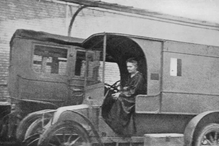 Marie Curie, a notable woman in history, sits in the driver's seat of an early model vehicle, possibly an ambulance.