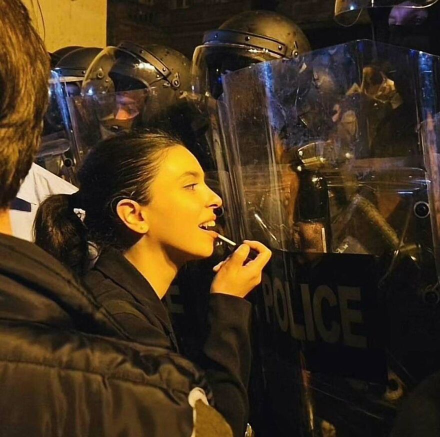 A woman applies lipstick in front of police officers with riot shields, a chaotic and disturbing pic.