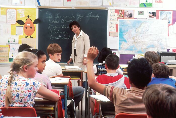 A teacher looking at students in a classroom, with one student raising their hand, revealing widely known industry secrets.