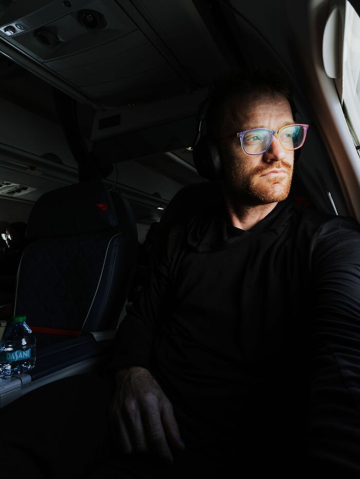 A man wearing headphones and glasses looking out of a plane window, contemplating entitled people.