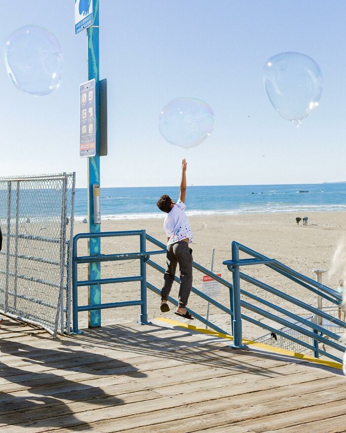 Perfectly timed street photo by Jeremy Paige of a boy reaching for large bubbles on a pier overlooking the ocean.