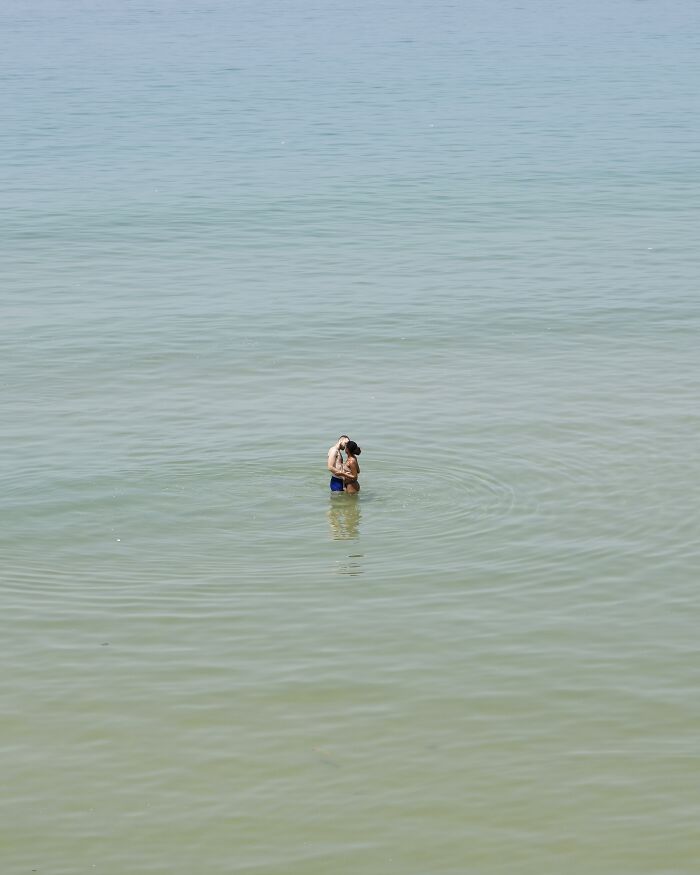 A photographer uses negative space to capture two people embracing in the vast, calm waters of a beach scene.