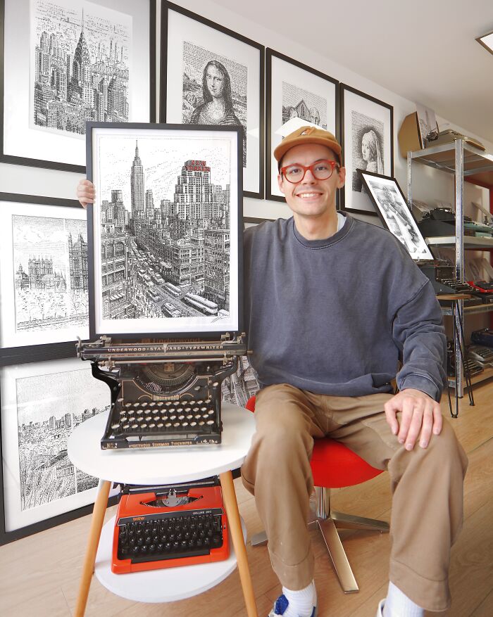 Artist smiling, holding a framed New York City typewriter art piece, with other art and typewriters visible.