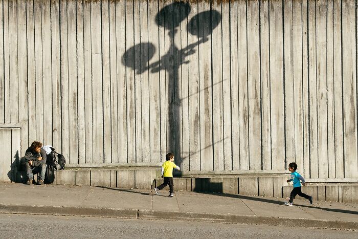Two children running on a sidewalk with a large shadow of a street lamp, a perfectly timed street photo by Jeremy Paige.