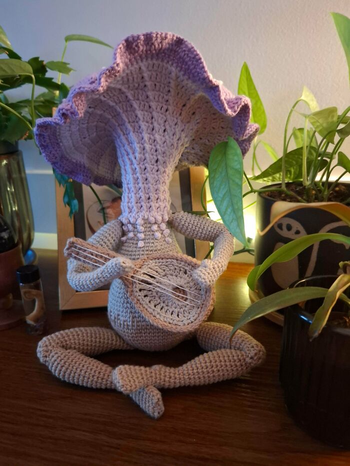Handmade crochet mushroom figure playing a stringed instrument surrounded by plants