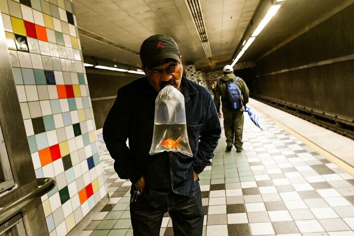 A man holds a bag with a goldfish in front of his face in a perfectly timed street photo by Jeremy Paige.