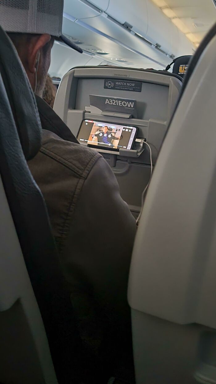 A passenger watching a video on a smartphone attached to the seatback, a common scene with entitled people on flights.