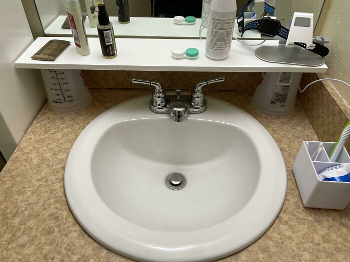 A DIY fail where a bathroom shelf is propped up by two plastic measuring cups above a sink.