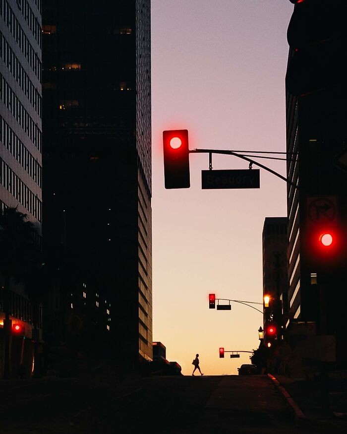 A perfectly timed street photo by Jeremy Paige of a silhouetted figure walking down a street at dusk with red traffic lights.
