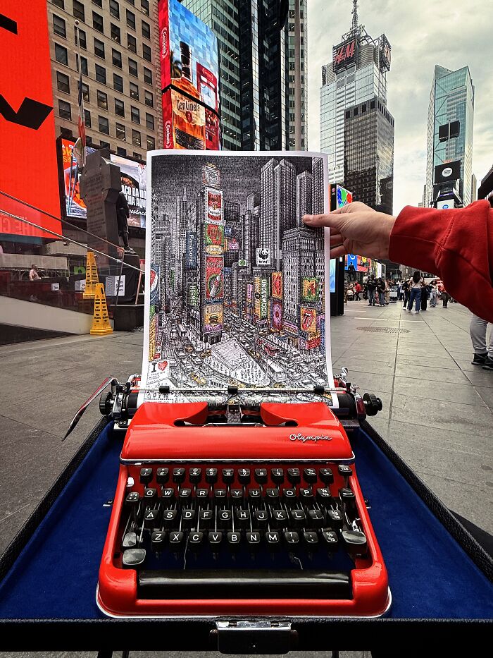 A red vintage typewriter on a blue surface, displaying a piece of Times Square typewriter art in a busy city environment.
