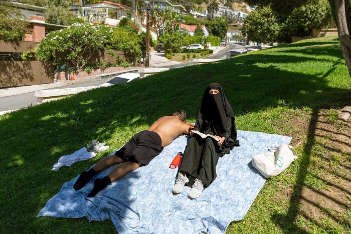 A person in a niqab reads a book while a man sunbathes on a picnic blanket, a perfectly timed street photo.