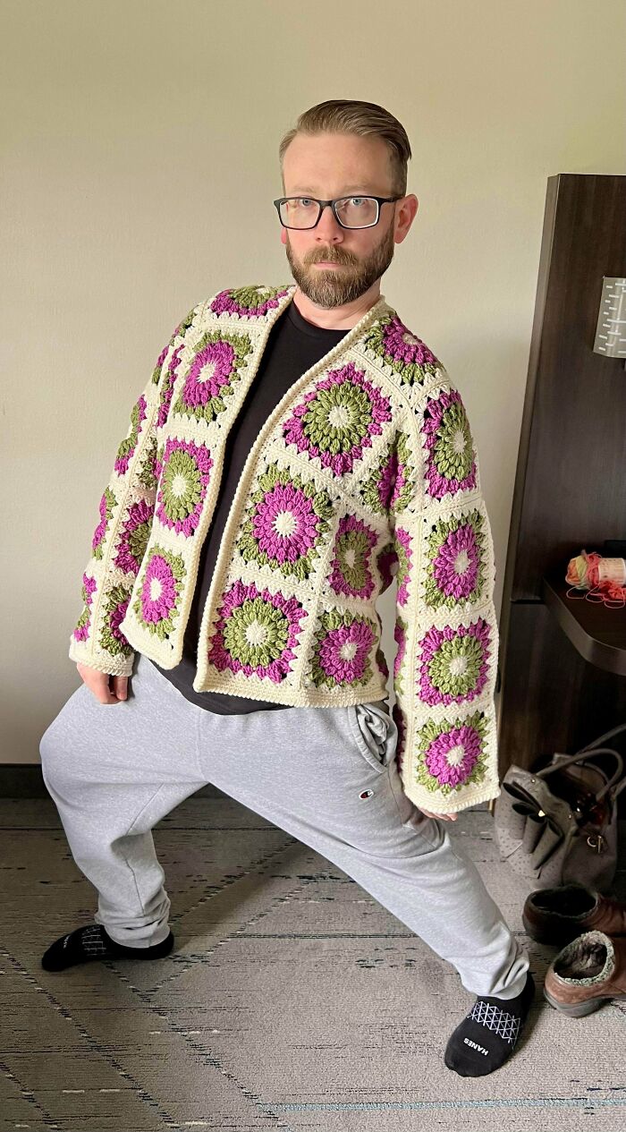 Man posing in a handmade crocheted cardigan with pink and green floral squares