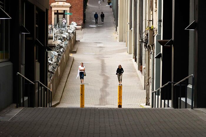 Two women stand on yellow bollards in a perfectly timed street photo by Jeremy Paige, looking like giants in a sloped alley.