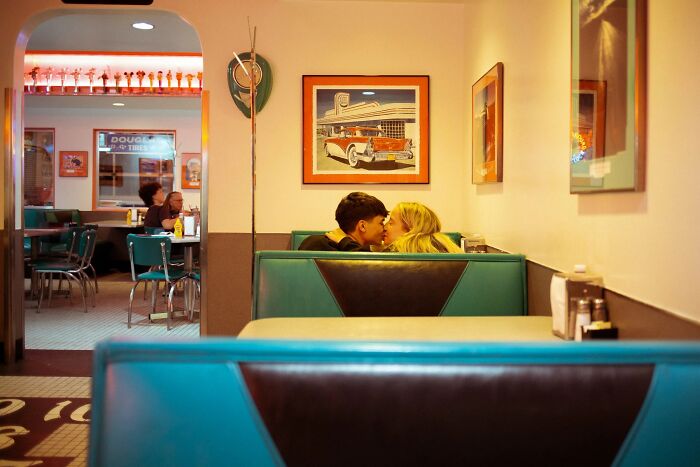 A couple shares a kiss in a diner booth, capturing a perfectly timed street photo moment by Jeremy Paige.