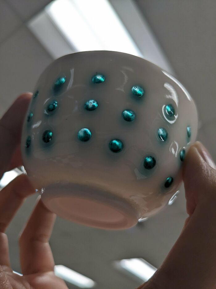 Hand holding a small white ceramic cup with raised teal dots, highlighting artistic pottery creations.