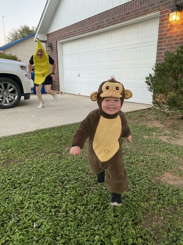 A toddler dressed in a monkey costume runs outside as an adult in a banana costume chases behind.