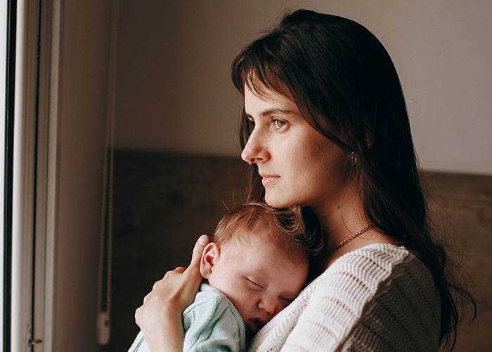 Mother thoughtfully holding her sleeping newborn baby near a window, reflecting on the realities of new motherhood