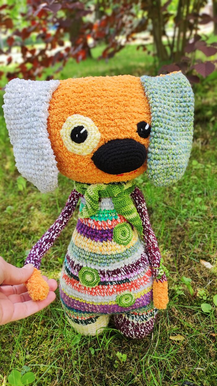 An adorable orange and grey dog-like crochet creature standing on green grass with a hand touching its arm