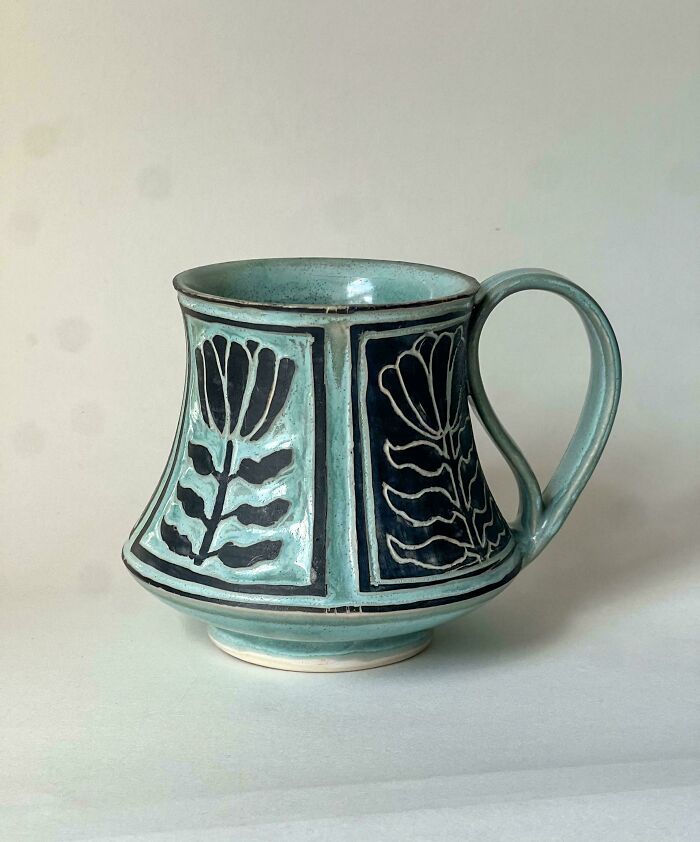 A light blue handmade pottery mug decorated with black floral patterns in relief.