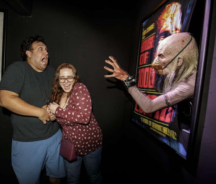 A man and woman scream in fear as a creature reaches out from a poster in a haunted house.