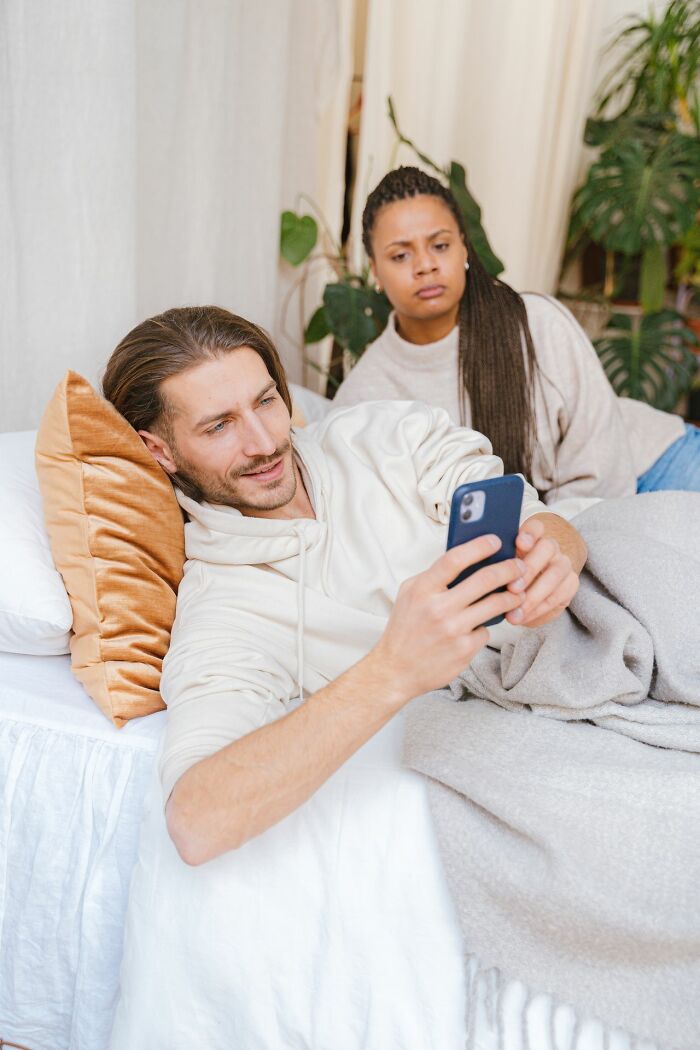 An annoyed woman watches her partner focus on his phone in bed, highlighting why marriages collapsed.