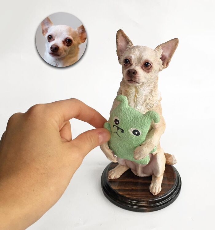 A human hand placing a toy in a hyperrealistic sculpture of a chihuahua sitting on a wooden stand