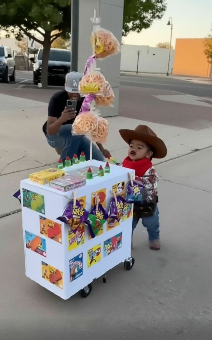 A toddler wearing a snack vendor Halloween costume pushing a white cart filled with chips and candy.
