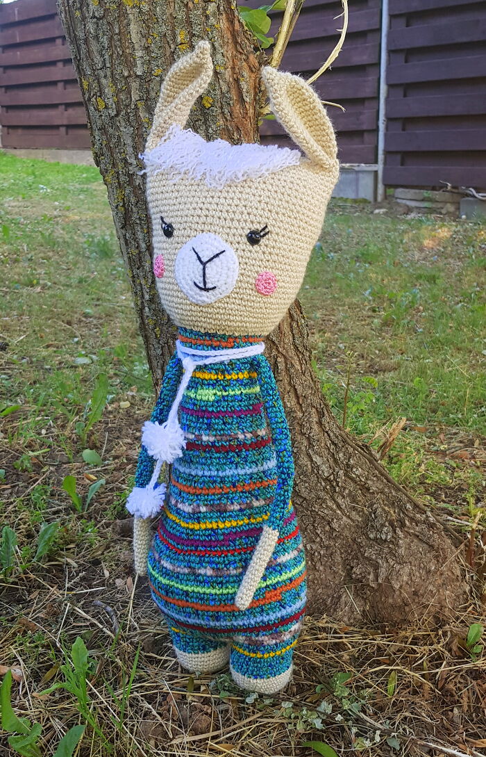 A handmade crochet creature llama leaning against a tree trunk in a grassy field