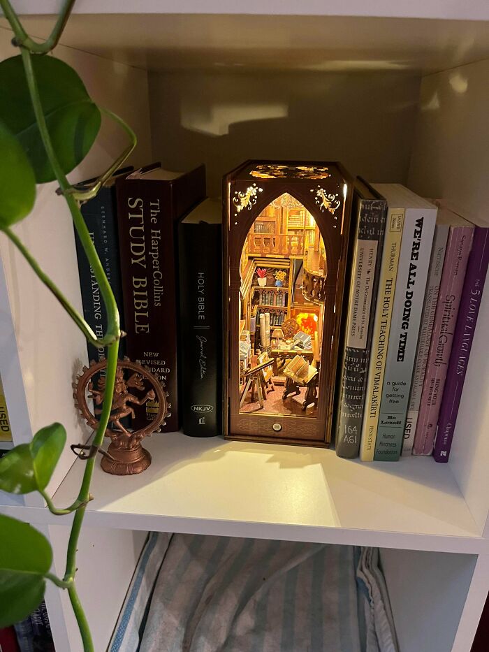 An illuminated wooden book nook shaped like an arched library nestled between real books on a white shelf.