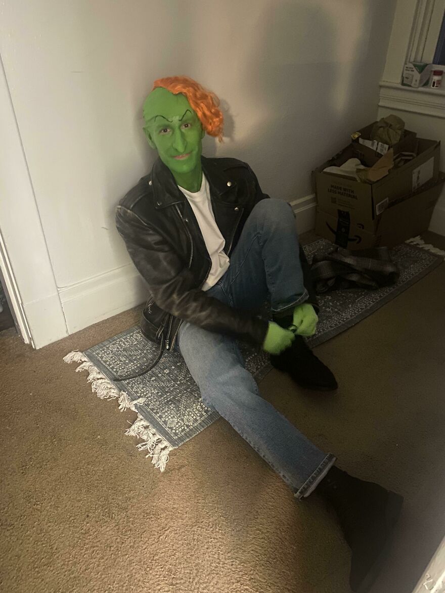 A person with green face paint and orange hair dressed as a goblin in a leather jacket for a Halloween costume idea.