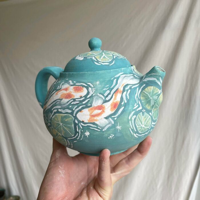 A hand holding a hand-painted light blue pottery teapot featuring orange koi fish and lily pads.