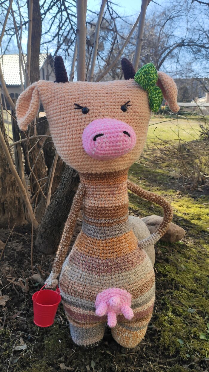 An orange striped crochet cow stands outdoors holding a small red bucket.
