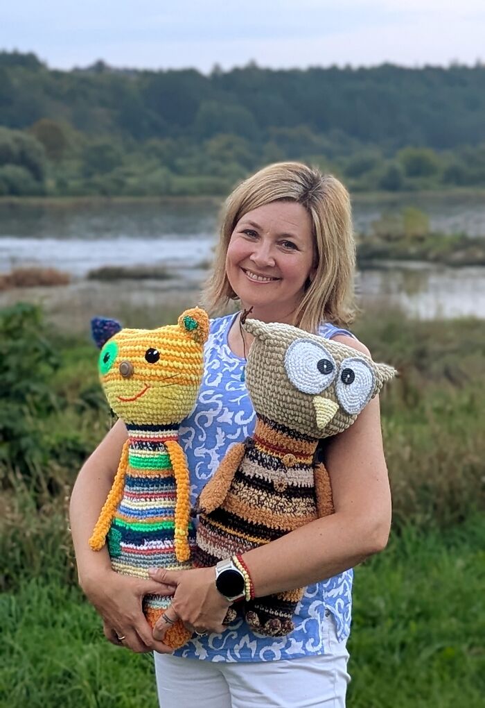 A woman happily holding two colorful crochet creatures, a striped cat and an owl, in front of a scenic river view