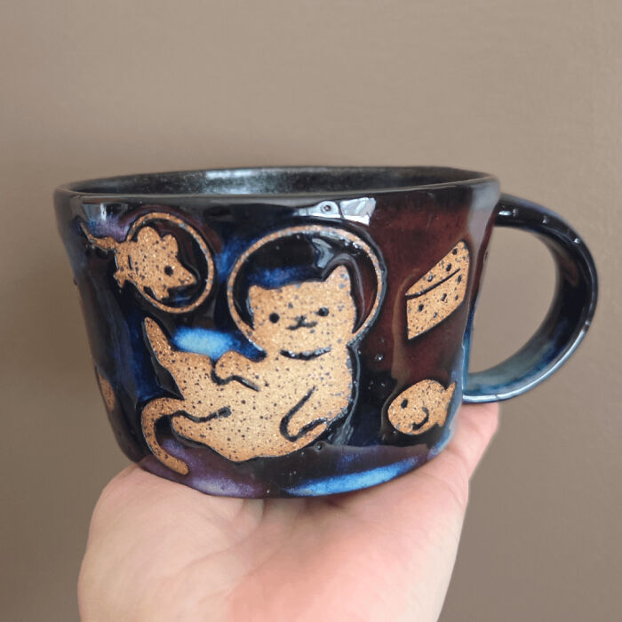 A dark glazed pottery mug decorated with a floating space cat and celestial details.
