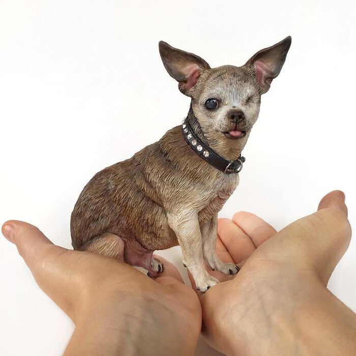 A hand holding one of the hyperrealistic sculptures of pets, featuring a winking Chihuahua wearing a collar.
