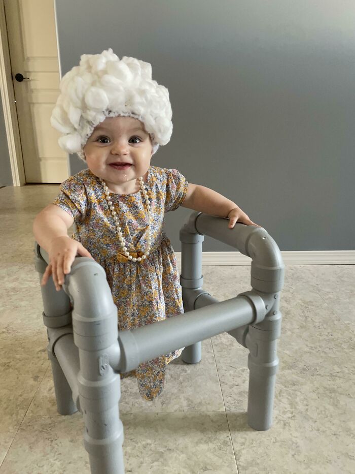 A baby wearing a grandmother costume for Halloween costumes with a white wig and walker
