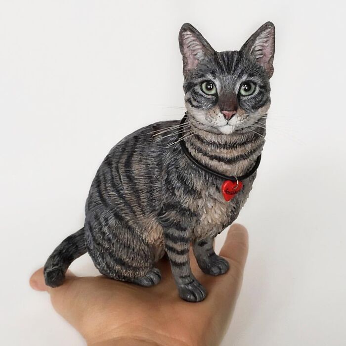 A realistic miniature grey tabby cat sculpture resting on an open palm, showcasing hyperrealistic sculptures of pets.