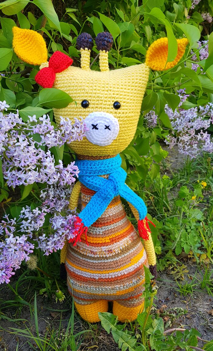 An adorable yellow crochet creature giraffe wearing a blue scarf and standing next to purple lilac flowers