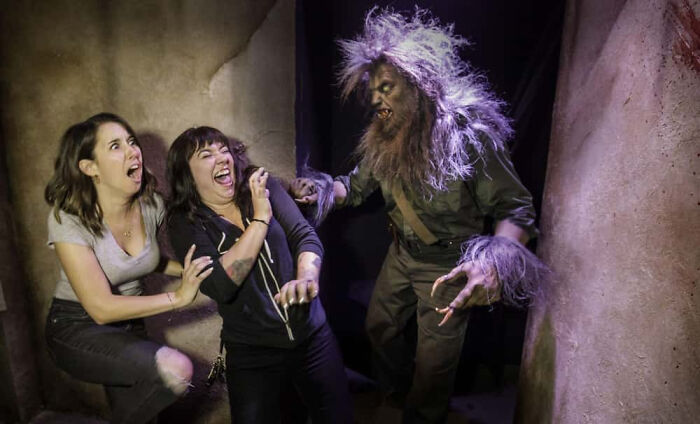 Two terrified women react with shock to a werewolf figure inside a haunted house.
